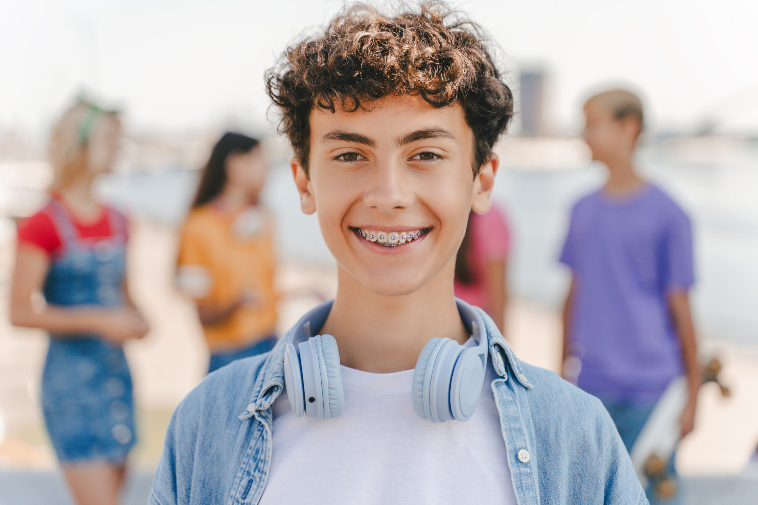 portrait of smiling teenage boy with braces wearing headphones looking at camera standing on the street with friends on background.