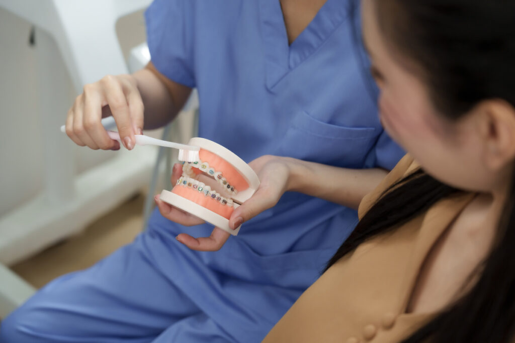 Asian female dental assistant demonstrating correct brushing technique on orthodontic model to adult patient. Explanation focused on oral hygiene and dental maintenance after braces treatment.