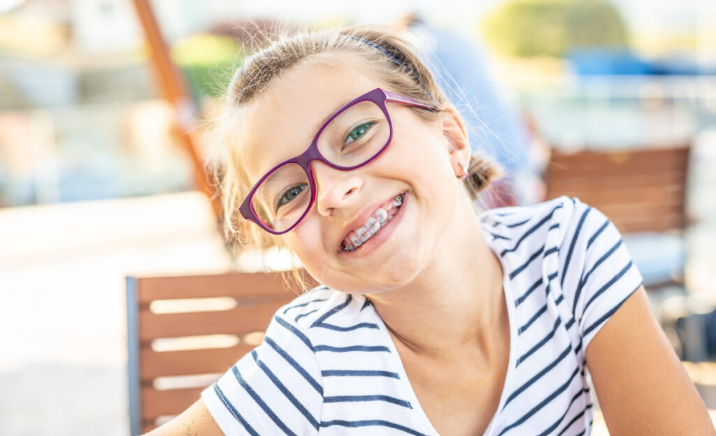 Young preteen girl in glasses wearing braces smiles at the camera on a summer day.