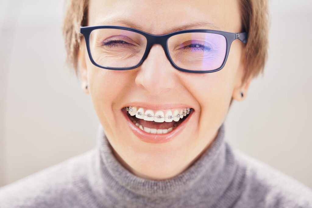 Stylish and beautiful young blonde woman in glasses and a gray sweater in braces and smiling on a white background, dentist orthodontist concept
