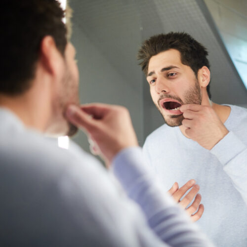 Handsome young man with stubble keeping mouth open while checking tooth and looking into mirror in bathroom